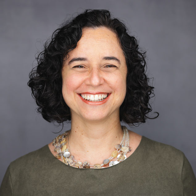 A professional headshot featuring a gray background with a smiling Brazilian woman with short, dark, curly hair and an olive green shirt.