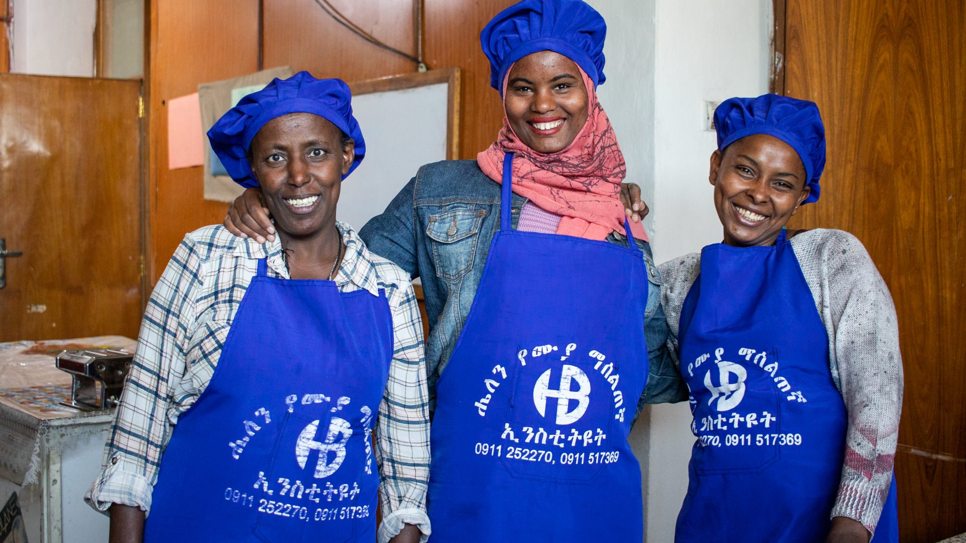 Three black women stand arm in arm, each wearing a royal blue apron and chef's hat.