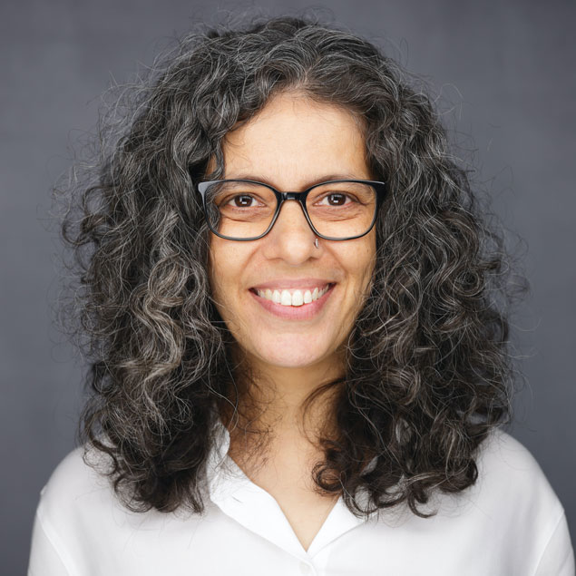 A professional headshot of a brown woman with shoulder length curly dark hair, a white blouse and glasses.