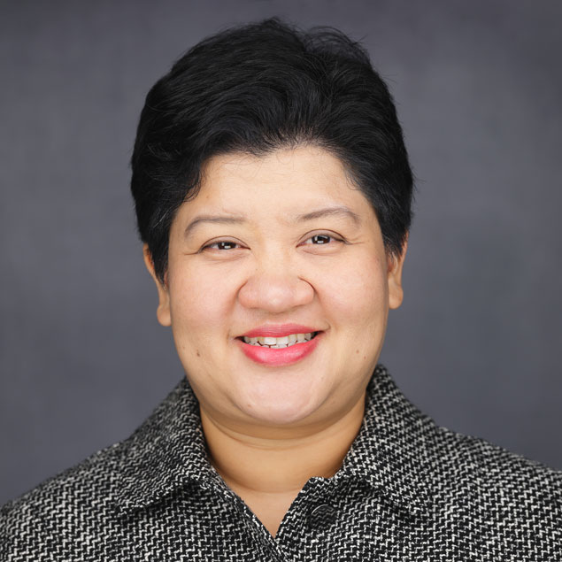 A professional headshot of a smiling Thai woman with short black hair and a collared shift, against a gray background.